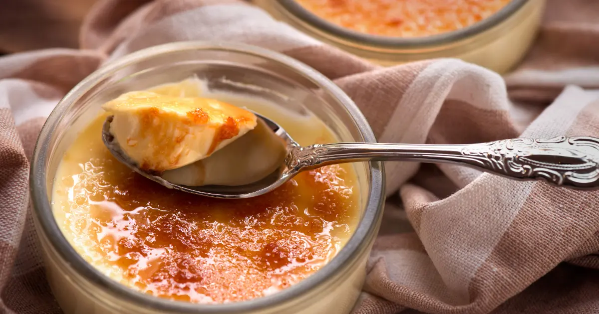 A photo of a crab brulee, showing a golden-brown caramelized sugar topping on a creamy, savory custard filled with chunks of tender crab meat, served in an elegant white ramekin on a dark slate background.