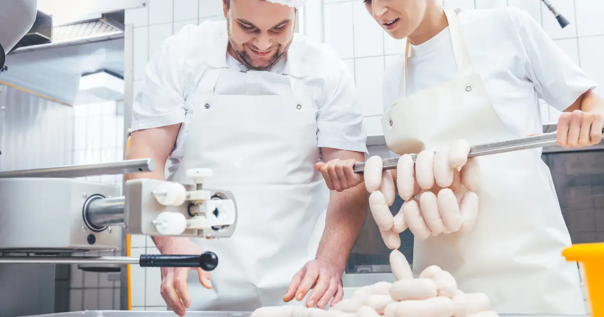 A man and a woman in a home kitchen, smiling as they prepare venison sausages together, focusing on choosing the right fat for their venison sausage, with various cooking ingredients spread out on the countertop