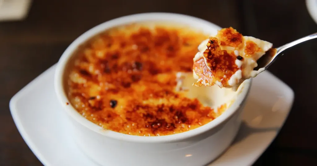 A close-up image of a freshly caramelized crème brûlée, showing a golden, crispy sugar crust that's lightly browned and cracked, revealing the smooth, creamy custard beneath.