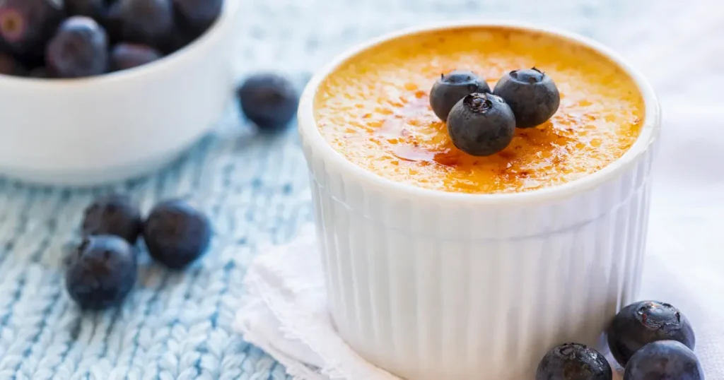 A close-up photo of a perfectly caramelized Crème Brûlée with a crispy sugar crust on top, served in a ramekin with blueberries on the top.