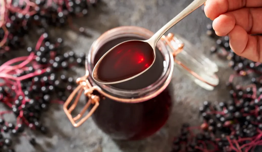 A jar of homemade elderberry syrup, with a spoon lifting some of the dark, translucent liquid, set against a backdrop of scattered elderberries and rustic textures.