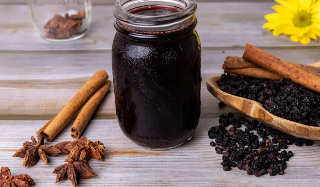 A glass jar filled with rich, dark elderberry syrup alongside a pile of dried elderberries, set against a neutral background.