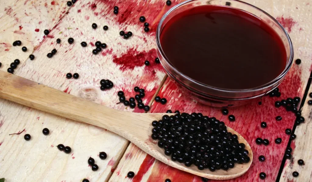 A spoon filled with fresh elderberries next to a jar of homemade elderberry syrup, displayed on a rustic wooden surface.