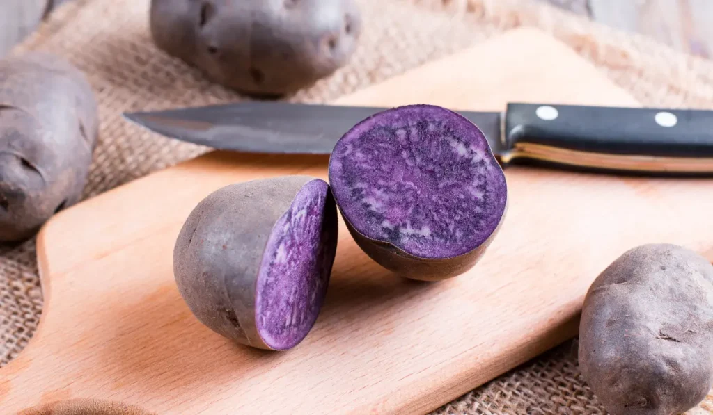 A sliced purple sweet potato on a wooden cutting board, showing its vibrant purple interior. Each slice is neatly arranged to display the cross-section, highlighting the smooth texture and intense color contrast between the purple flesh and the darker skin.